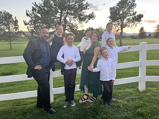 A family dressed in semi-formal attire posing together by a white fence in a park setting with trees and a clear sky in the background.